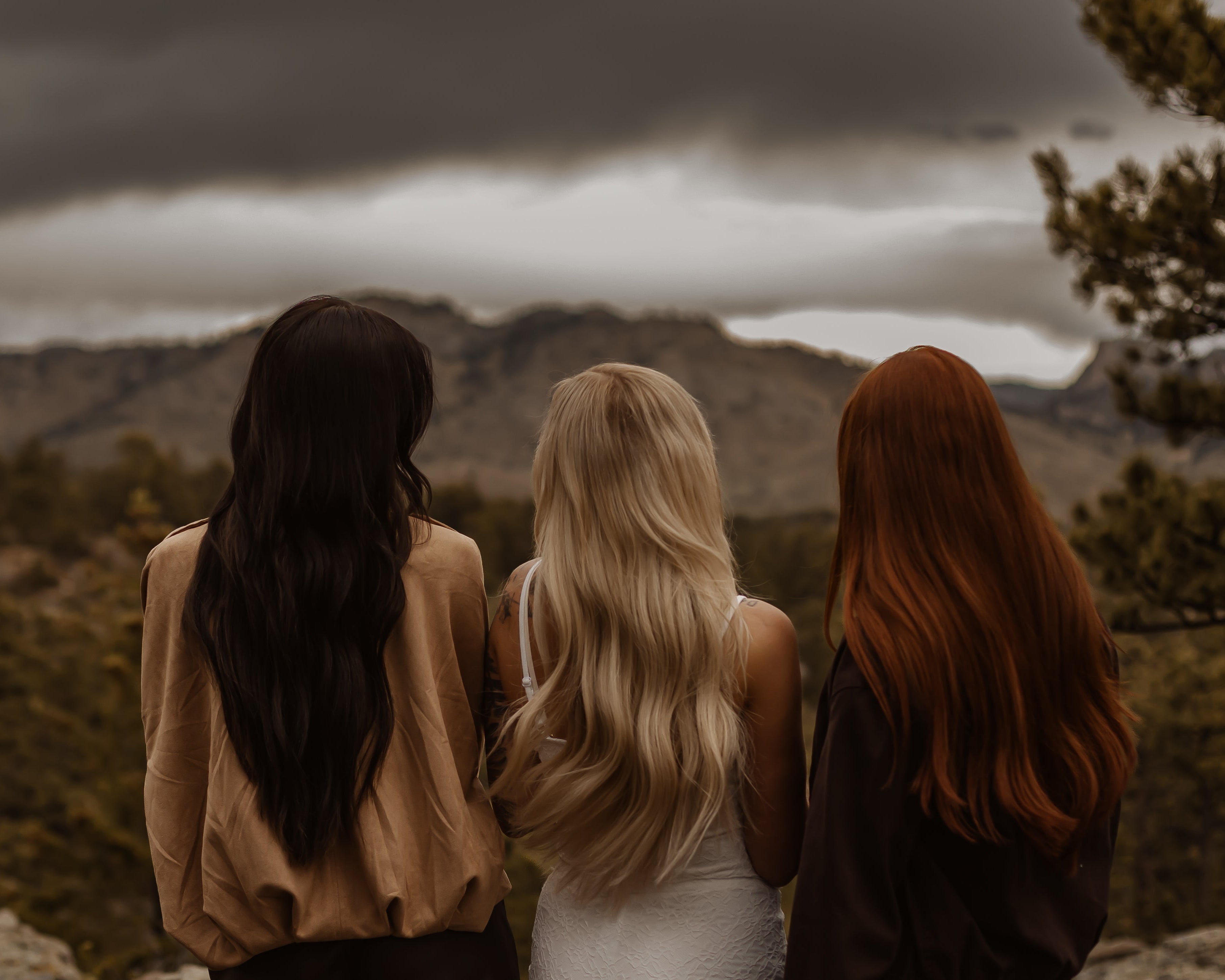 Three women seen from behind standing outdoors in a mountain landscape under a dark cloudy sky. The woman on the left has long black hair and wears a tan top, the center woman has long blonde hair and wears a white sleeveless top, and the woman on the right has long red hair and wears a dark brown top.