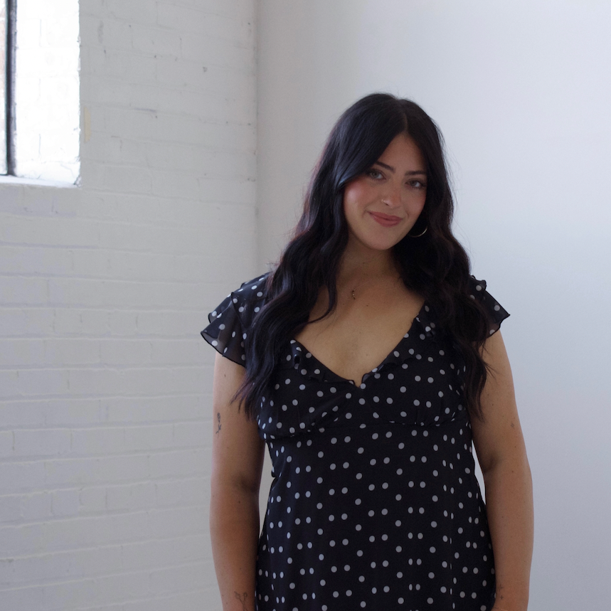 A woman with long dark hair wearing a black V-neck dress with white polka dots, standing in a bright room with white brick walls.