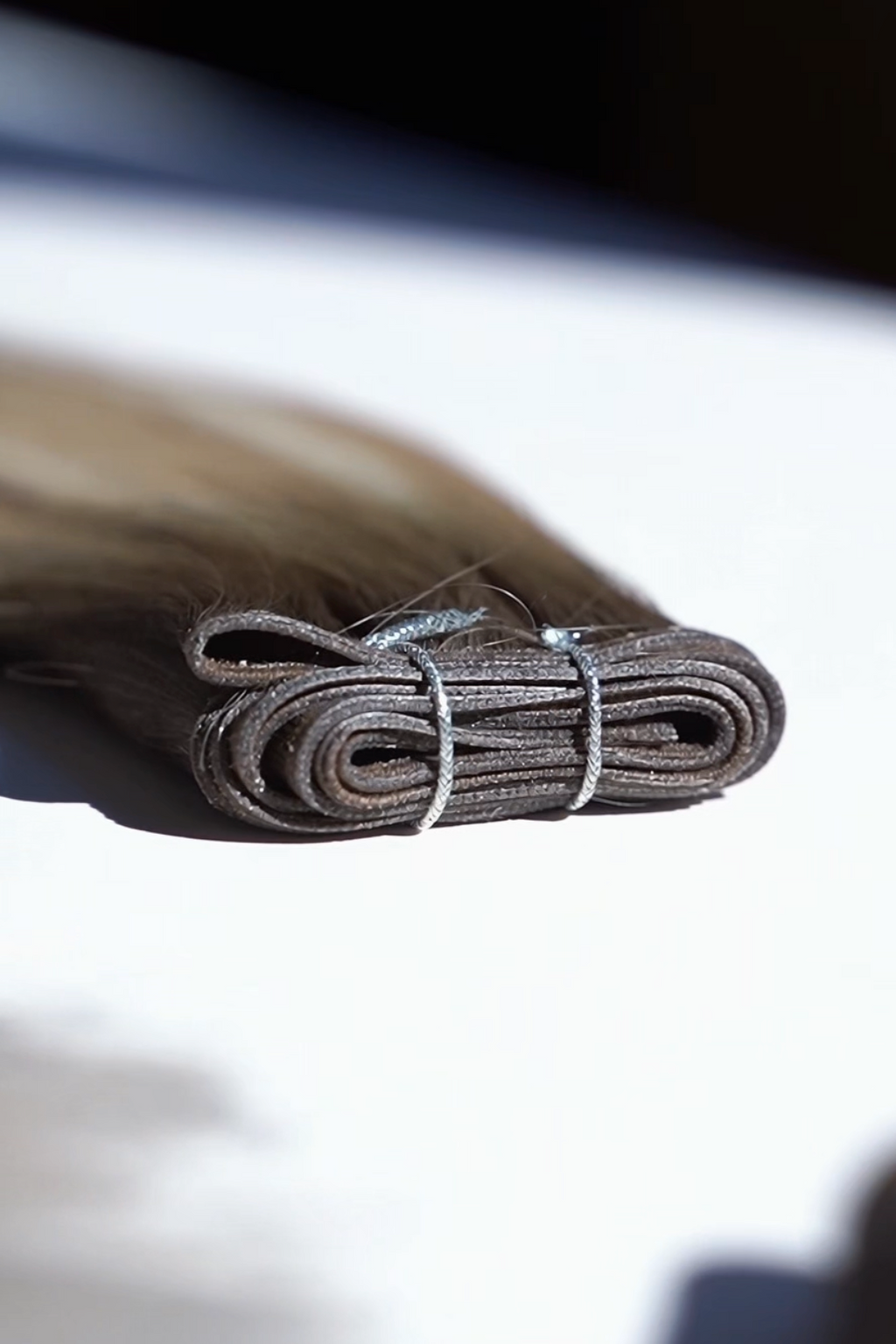 A close-up of a bundled bundle of light brown hair extensions tied with a silver wire, resting on a white surface with a dark, blurred background.