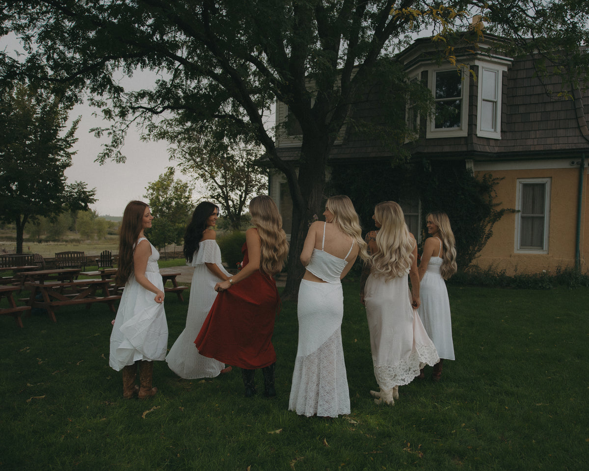 Six women wearing white and red dresses stand in a circle on a grassy lawn in front of a large tree and a house, viewed from behind.
